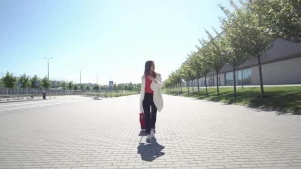 Mulher bonita na camisa vermelha caminha com mala fora em um dia ensolarado — Vídeo de Stock