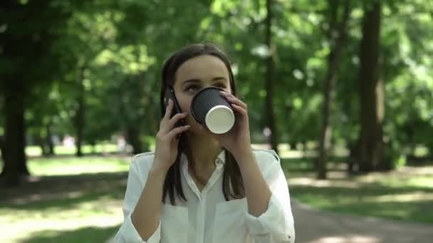 Mulher bebe café e fala no telefone no parque — Vídeo de Stock