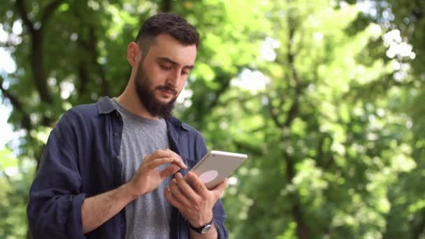 El hombre trabaja con la tableta de pie en el parque — Vídeo de stock