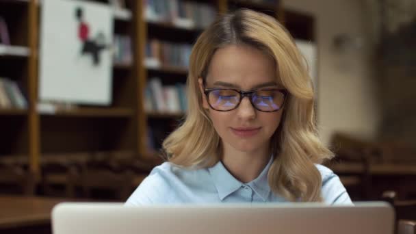Блондинка працює з ноутбуком у бібліотеці — стокове відео
