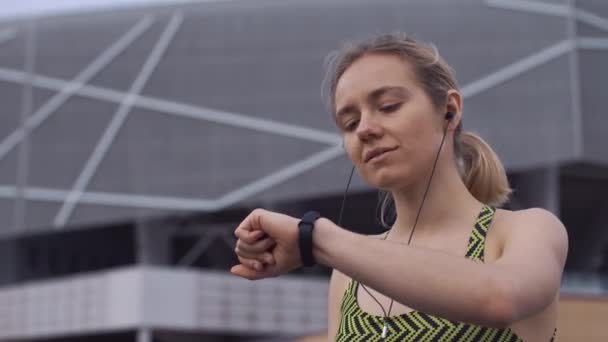 Женщина в спортивном костюме слушает музыку, сидящую на скамейке и проверяет свой фитнес-браслет — стоковое видео
