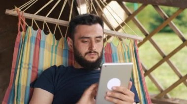 Bir ev bir hamakta yatan genç sakallı biri bir tablet kullanır