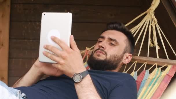 Egy fiatal szakállas ember használ egy tabletta, fekszik egy függőágyban, egy vidéki házban — Stock videók