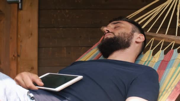Ένας νεαρός γενειοφόρος άνδρας που κοιμούνται σε μια αιώρα σε μια εξοχική κατοικία. Κοιμάται με tablet — Αρχείο Βίντεο