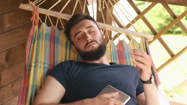 一个年轻的有胡子的人睡在吊床挂在乡间别墅。他着平板电脑睡觉 — 图库视频影像