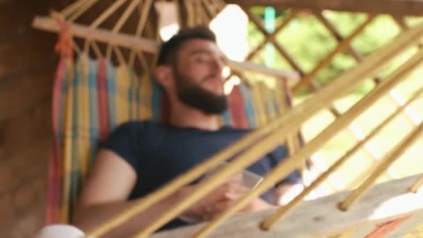 Un joven barbudo durmiendo en una hamaca en una casa de campo. Duerme con la tableta — Vídeo de stock