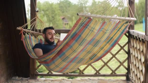 Un jeune homme barbu utilise une tablette, communique par vidéoconférence, couché dans un hamac dans une maison de campagne — Video
