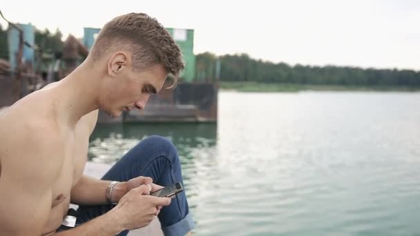 Чоловік сидить на бухті зі своїм телефоном — стокове відео