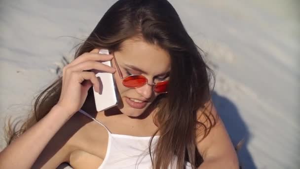 Verleidelijke brunette in rode zonnebril gesprekken over de telefoon liggend op het strand — Stockvideo