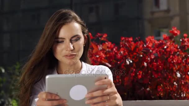 Брюнетка работает с планшетом, пока сидит в ресторане перед красными цветами — стоковое видео
