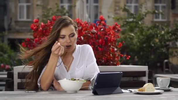 Brunette dame spiser salat og ser noget på hendes tablet, mens hun sidder i restauranten før røde blomster – Stock-video