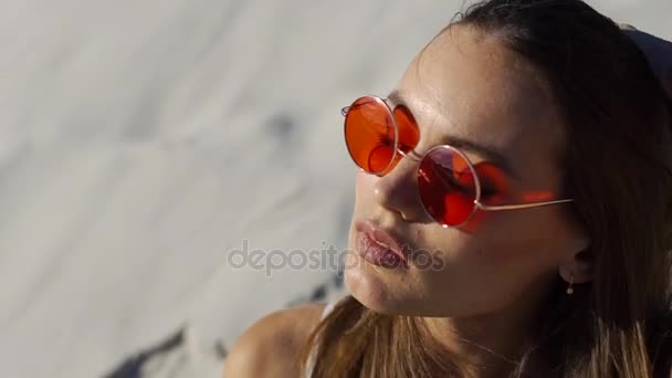 Mujer con gafas de sol rojas sacude su cabello sentado en la arena en un día soleado — Vídeos de Stock