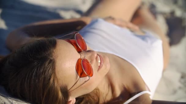 Спокуслива брюнетка в червоних сонцезахисних окулярах лежить в нижній білизні на піску — стокове відео