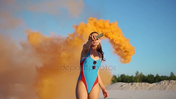 Brunette woman in blue swimming suit plays with orange smoke on the beach — Stock Video