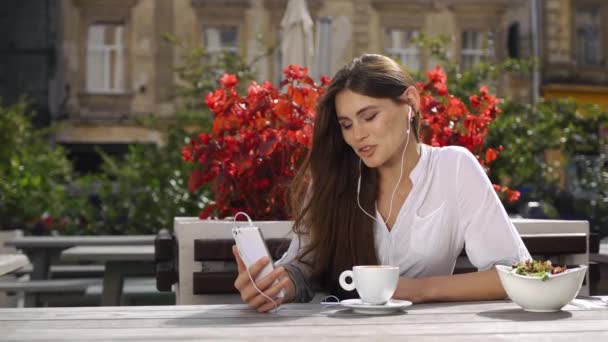 Morena dama habla en su teléfono a través de auriculares mientras se sienta en el restaurante antes de las flores rojas — Vídeos de Stock