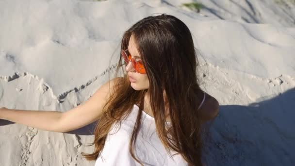 Mujer morena seductora en top blanco y gafas de sol rojas juega con la arena en el viento — Vídeos de Stock