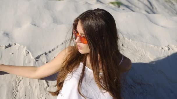 Femme brune séduisante en haut blanc et lunettes de soleil rouges joue avec le sable sur le vent — Video