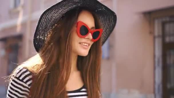 Vrouw in zwarte hoed en rode zonnebril glimlacht wervelende op straat — Stockvideo