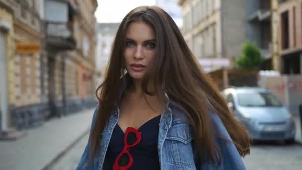 Mujer morena con el pelo largo sonríe y gira caminando por la calle en su chaqueta jeans — Vídeos de Stock