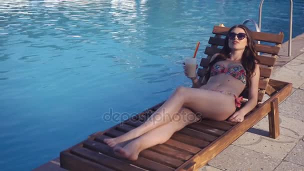 美丽的年轻女子，穿着比基尼喝鸡尾酒、 坐在泳池边和日光浴 — 图库视频影像