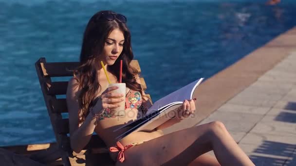 Jovem mulher bonita lendo o livro e beber um coquetel perto da piscina — Vídeo de Stock