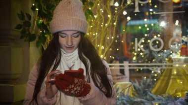Güzel çekici kız bir window-shop ayakta çelenk ile dekore edilmiş ve bir akıllı telefon kullanır. Yeni yıl Noel. Akşam çekim