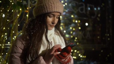 Güzel çekici kız bir window-shop ayakta çelenk ile dekore edilmiş ve bir akıllı telefon kullanır. Yeni yıl Noel. Akşam çekim