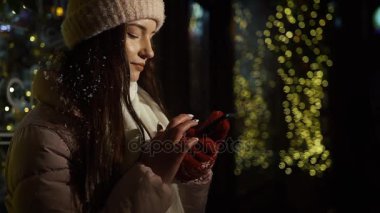 Güzel çekici kız bir window-shop ayakta çelenk ile dekore edilmiş ve bir akıllı telefon kullanır. Yeni yıl Noel. Akşam çekim