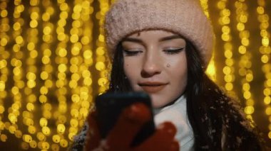 Güzel çekici kız bir window-shop ayakta çelenk ile dekore edilmiş ve bir akıllı telefon kullanır. Yeni yıl Noel. Akşam çekim