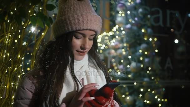 Hermosa chica atractiva de pie cerca de un escaparate decorado con guirnaldas y utiliza un teléfono inteligente. Año Nuevo Navidad. Disparos nocturnos — Vídeos de Stock