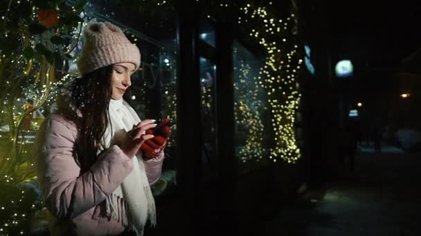 Menina bonita atraente de pé perto de uma vitrine decorada com guirlandas e usa um smartphone. Natal de Ano Novo. Disparo nocturno — Vídeo de Stock