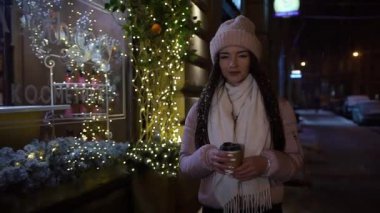 Onun elinde kahvesiyle çelenk süslenmiş ve kameraya bakarak vitrinler, yakınındaki sokakta yürürken güzel çekici kız. Yeni yıl Noel. Akşam çekim