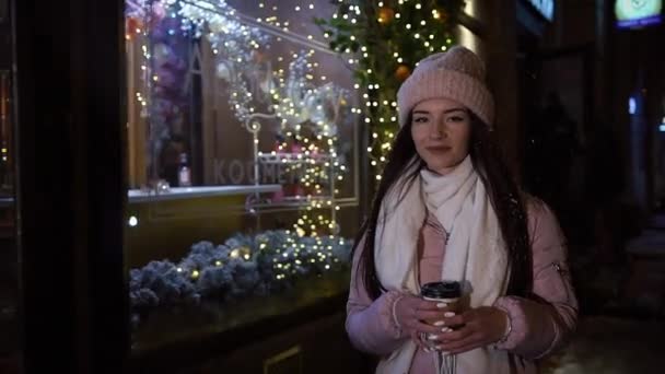 Menina bonita e atraente andando pela rua perto de vitrines, decorada com guirlandas, com café nas mãos e olhando para a câmera. Natal de Ano Novo. Disparo nocturno — Vídeo de Stock