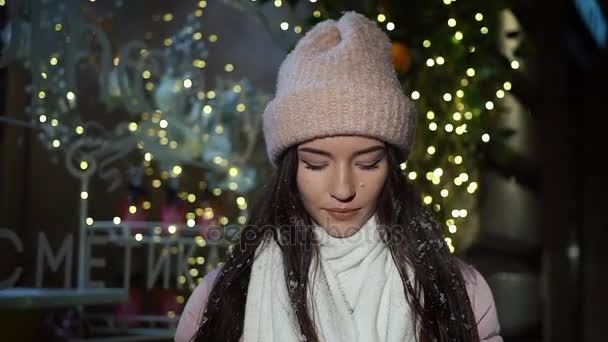 美丽迷人的女孩站在一个装饰着圣诞彩灯和看着相机的橱窗商店旁边。咖啡在女孩的手里。她喝咖啡和微笑。特写, 夜间拍摄 — 图库视频影像