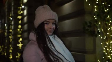 Çelenk ile dekore edilmiş ve kameraya bakarak vitrinler, yakınındaki sokakta yürürken güzel çekici kız. Yeni yıl Noel. Akşam çekim