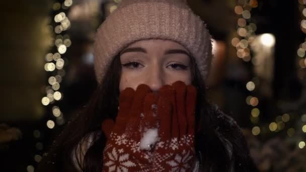 Bella ragazza attraente sta camminando intorno alla sera accanto a un negozio di vetrine decorate con luci di Natale e guardando nella fotocamera. Lancia un bacio aereo. Rallentatore, Primo piano — Video Stock