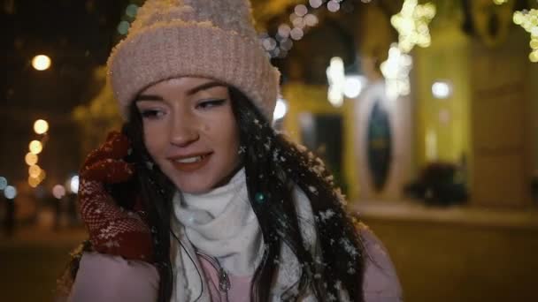 Gyönyörű, vonzó lány az utcán közelében kirakatok, díszített koszorúkat, és beszél a telefonon. Új év karácsony. Este lövés közelről — Stock videók