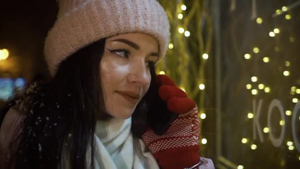 Όμορφη ελκυστικό κορίτσι στέκεται κοντά ένα window-shop διακοσμημένα με γιρλάντες και και μιλάει μέσω τηλεφώνου. Χριστούγεννα Πρωτοχρονιά. Βράδυ, σκοποβολή, γκρο πλαν — Αρχείο Βίντεο