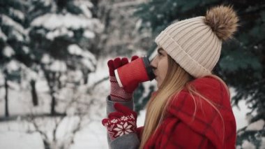 Genç güzel kız içinde Noel ağaçları karda sıcak içecek kağıt bardak içme yakınındaki sıcak giysiler standinh. Ağır çekim. Bir kış parkta şık genç güzel kız portresi