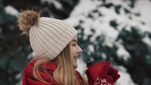 Giovane bella ragazza in abiti caldi in piedi vicino agli alberi di Natale nella neve bere bevanda calda dal bicchiere di carta. Al rallentatore. Primo piano — Video Stock