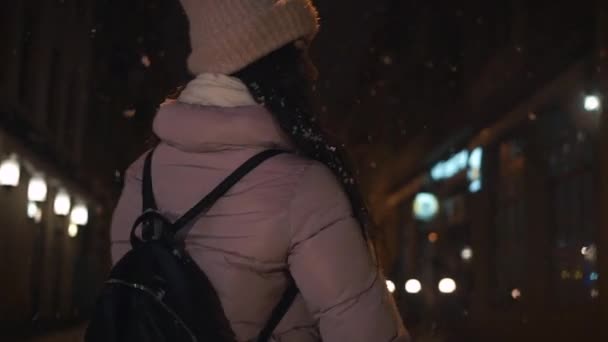 美しい魅力的な女の子は、夕方には雪に覆われた天気の都市の通りを通って行きます。少女は夜の街で歩いています。スローモーション、降雪 — ストック動画