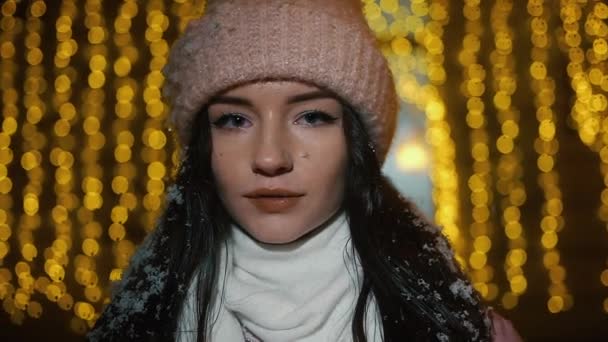 Beautiful attractive girl standing near a window-shop decorated with garlands and and looking into the camera. New Year Christmas. Evening shooting, Slow motion, snowfall — Stock Video