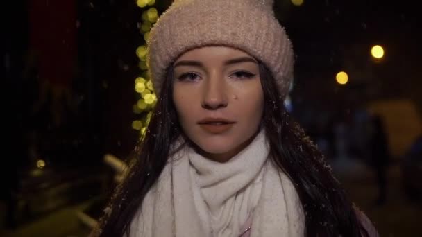 Menina bonita atraente em pé perto de uma vitrine decorada com guirlandas e olhando para a câmera. Natal de Ano Novo. Tiro à noite, câmera lenta, queda de neve — Vídeo de Stock