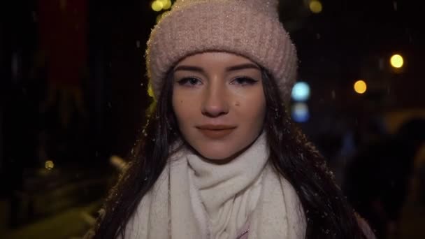 Bella ragazza attraente in piedi vicino a un negozio di finestre decorato con ghirlande e guardando nella fotocamera. Natale di Capodanno. Scatto serale, rallentamento, nevicate — Video Stock