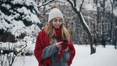 Noel ağaçları duran sıcak giysiler içindeki genç güzel kız ve ve bir akıllı telefon kullanır. Ağır çekim. Bir kış parkta şık genç güzel kız portresi