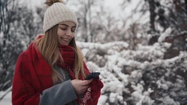 Joven chica hermosa en ropa de abrigo de pie cerca de los árboles de Navidad y y utiliza un teléfono inteligente. En cámara lenta. Retrato de la joven y elegante hermosa chica en un parque de invierno — Vídeos de Stock