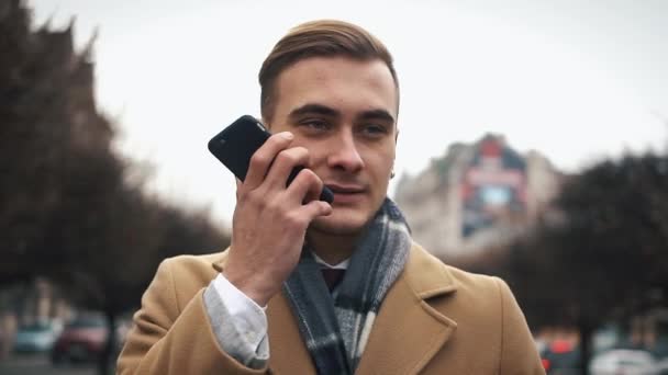 Un joven hombre de negocios en una capa caminando por la calle en un clima nublado y hablando por teléfono — Vídeos de Stock