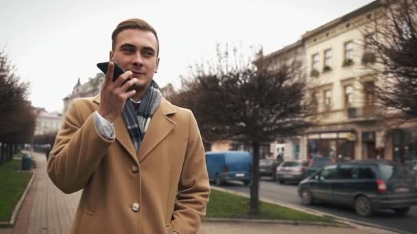 Un jeune homme d'affaires dans un manteau marchant dans la rue par un temps nuageux et parlant au téléphone — Video