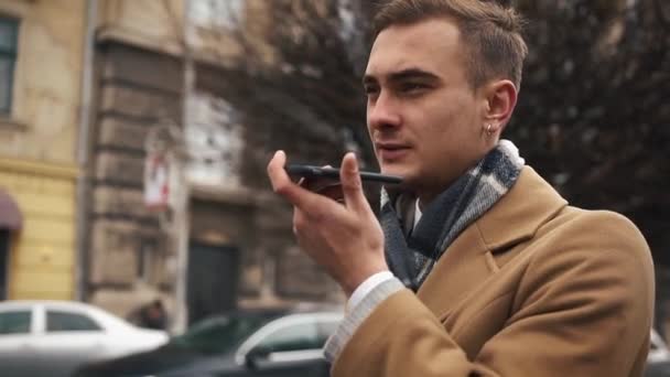 Молодой бизнесмен в плаще идет по улице в облачную погоду и разговаривает по телефону — стоковое видео