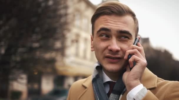 Ένας νεαρός επιχειρηματίας σε ένα μανδύα περπάτημα στο δρόμο με ένα ΣΥΝΝΕΦΙΑ και μιλάει στο τηλέφωνο. Αργή κίνηση — Αρχείο Βίντεο